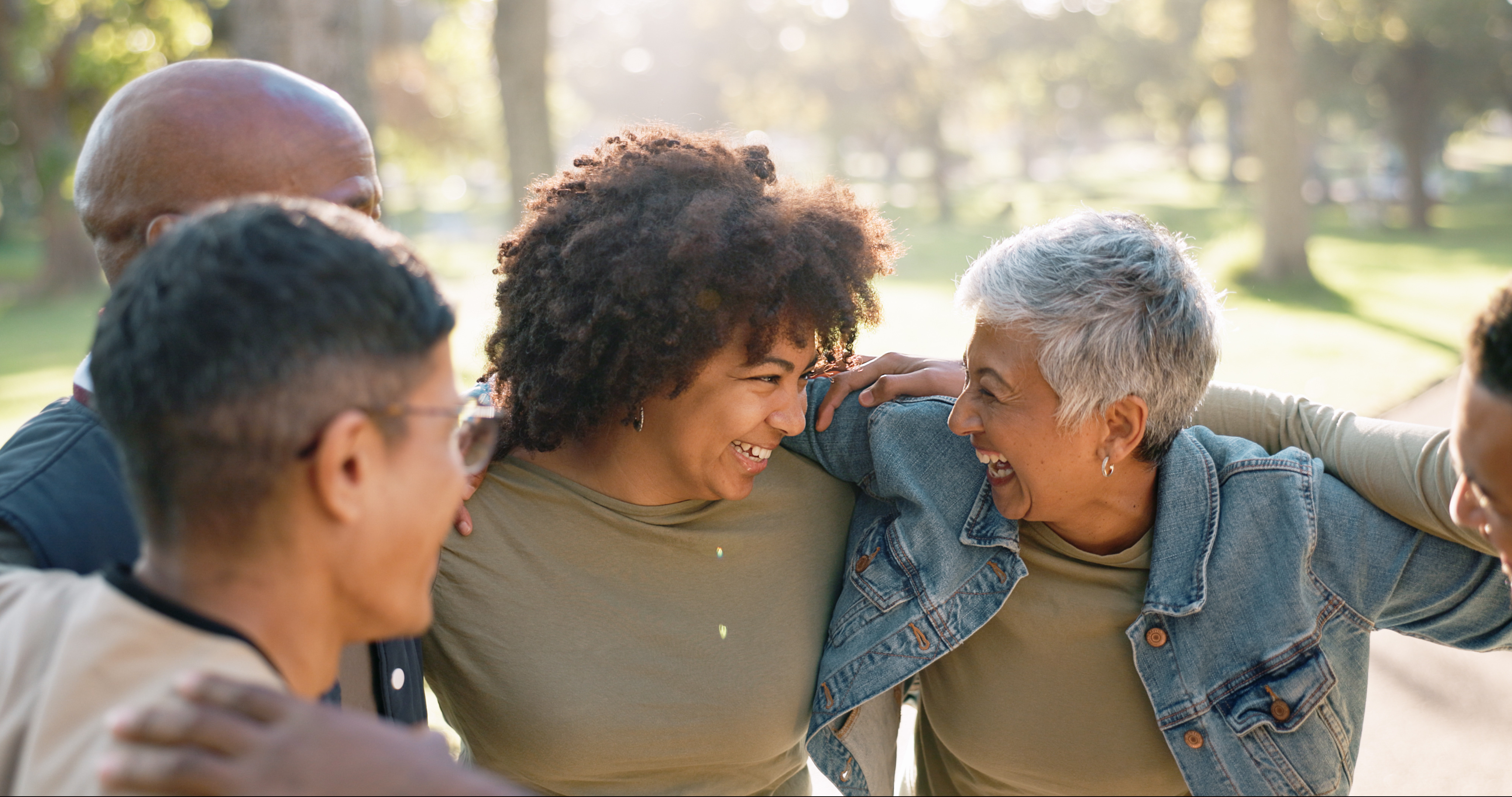 Diverse friends laugh together outdoors in a sunlit park, embracing with arms around one another and sharing a warm, joyful moment